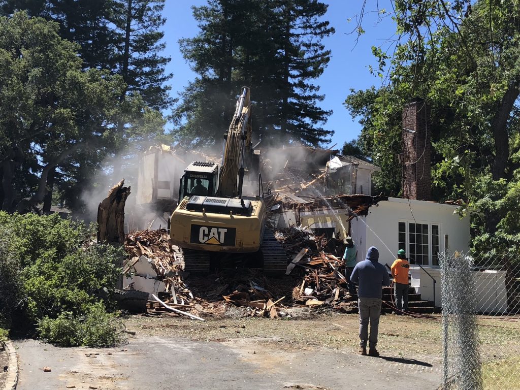 Excavator demolishing a damaged house with workers on site, showcasing the critical role of demolition companies in disaster recovery and cleanup.
