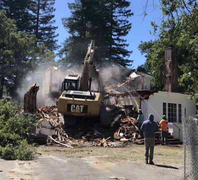 Excavator demolishing a damaged house with workers on site, showcasing the critical role of demolition companies in disaster recovery and cleanup.